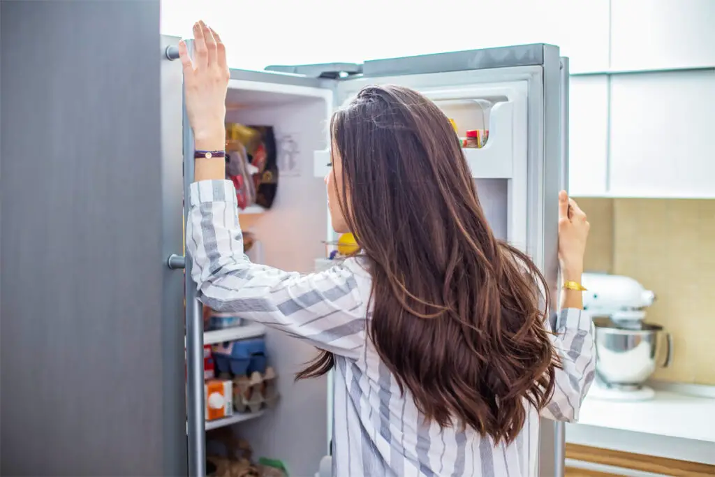 Frau steht vor Kühlschrank und schaut in den Kühlschrank. Für wie lange bleibt Wurst im Kühlschrank frisch?