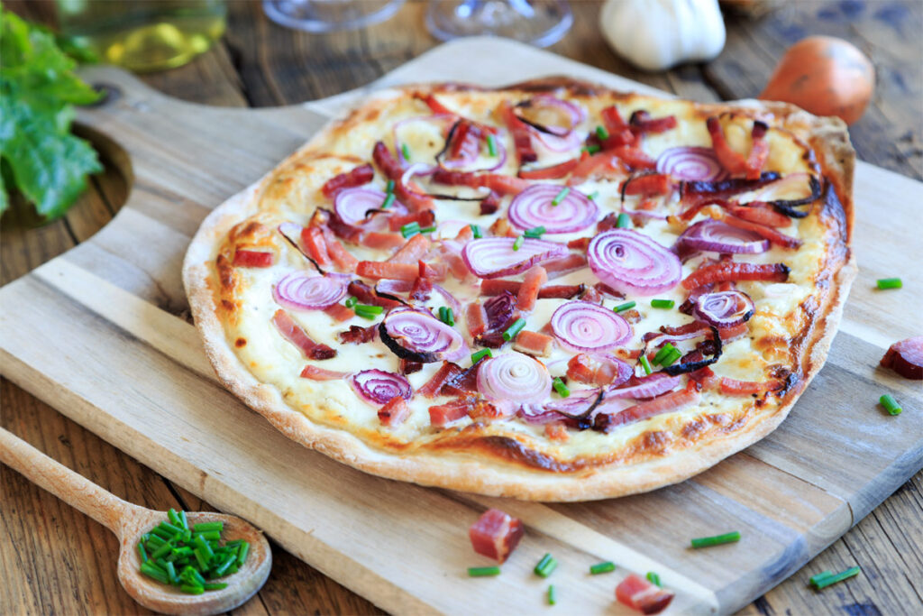 Flammkuchen mit Zwiebeln auf einem Holzbrett mit einem danebenliegenden Holzlöffel